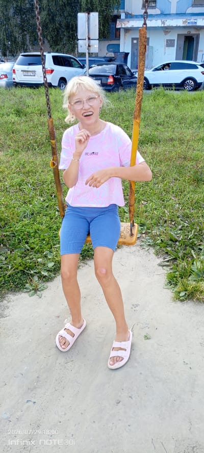 A young Caucasian female with blonde hair, glasses, and a pink and white tie-dye t-shirt sits on a yellow swing set in a park in Langepas, Russia. She is wearing blue athletic shorts and pink slides. Her left knee has a light-colored bandage or scar. The background shows green grass, trees, and buildings, as well as several parked cars. The overall scene suggests a casual outdoor setting on a daytime. The timestamp on the image indicates July 29, 2026, at 18:00.