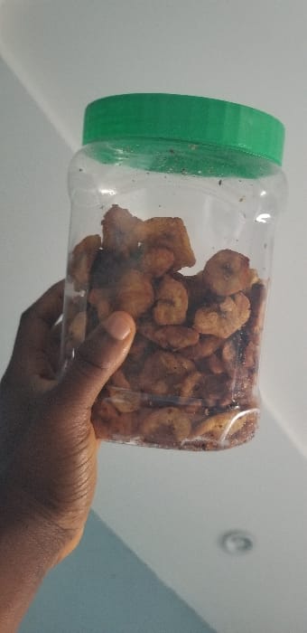 A dark-skinned right hand is observed gripping a clear, cylindrical plastic jar approximately three-quarters full of golden-brown, irregularly shaped fried plantain chips (kpekere). The jar is sealed with a solid green screw-top lid. Dark particulate matter, consistent with seasoning, is visible on the chips and adhered to the interior surface of the container. The background consists of an indoor environment, featuring a light-colored, possibly off-white, ceiling with a partially visible circular recessed light fixture in the upper right quadrant of the image. The lower portion of the background suggests a light-colored wall. This scene is located in the city of Ado-Ekiti, Nigeria.