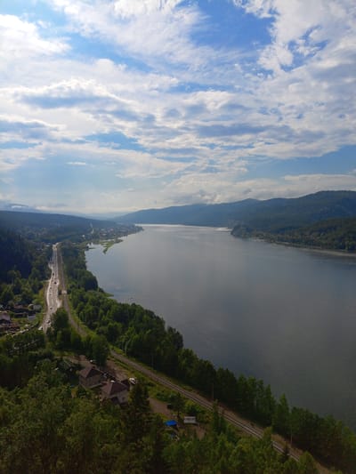 This aerial photograph captures a serene landscape of a wide, calm river winding through a valley. The river dominates the right side of the image, reflecting the cloudy blue sky. On the left side of the river, a lush, green hillside is covered in dense trees. A road runs parallel to the river, with a few cars visible. Adjacent to the road are train tracks.
Further inland, a small town or village is nestled along the riverbank, with houses and buildings clustered together. The scene is bathed in daylight, likely mid-morning or early afternoon, given the brightness and the presence of scattered clouds in the sky. The weather appears pleasant, with a mix of sun and clouds creating a gentle ambiance.
In the foreground, closer to the viewer, are more houses and trees, indicating a residential area on the hillside overlooking the river. The overall impression is one of peace and natural beauty.
There is a visible sign in Cyrillic script near the train tracks that reads "СЛИЗНЕВО". This suggests the location is Sliznevo, Russia. The image evokes a sense of tranquility and the vastness of nature.