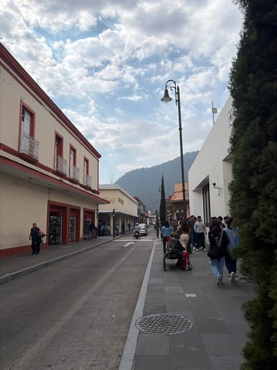 A street scene with people lining the sidewalk. Buildings flank the street, one with a cream facade and red trim. A street lamp stands tall against a partly cloudy sky. A mountain rises in the background. Cars and a motorcycle travel on the road, adding to the urban activity.