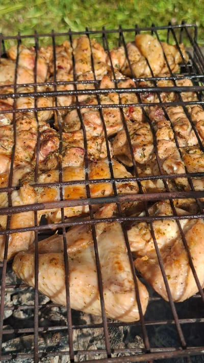 The image shows chicken pieces cooking on a metal grill, likely outdoors. The grill sits over what appears to be a charcoal or wood fire. The chicken is seasoned with herbs and spices. Part of a grassy area is visible in the background. The scene likely takes place in Сваткаўскі сельскі Савет, Belarus.