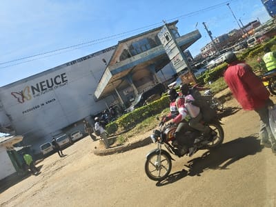 An outdoor, tilted photograph depicts a daytime street scene in Kapsabet, Kenya. The immediate foreground shows an unpaved road surface with prominent shadows indicating strong sunlight from a clear blue sky.
In the midground, a dark-colored motorcycle is visible with two male occupants. The driver wears a dark helmet, a red and white long-sleeved shirt, and a reflective vest. The male passenger, also wearing a reflective vest, carries a light-colored bag on his lap. To the right of the motorcycle, a male individual in a red jacket and dark pants stands, holding a light-colored bag, with his back partially to the camera. A second individual wearing a green reflective vest is partially visible on the far right edge of the frame.
On the left, a large, light-colored building is labeled "KIRSIBAK BUILDING" at the top and features prominent "NEUCE THE FUTURE OF PAINT" and "PRODUCED IN KENYA" signage on its side. Several individuals are visible near the building's entrance, and at least three white cars are parked in front of it. Adjacent to this, to the right, a multi-story structure labeled "SASA SERVICE STATION" is present, featuring a distinct covered area. Green shrubbery lines a raised median separating the unpaved road from the buildings. Overhead utility lines are visible across the upper portion of the frame.