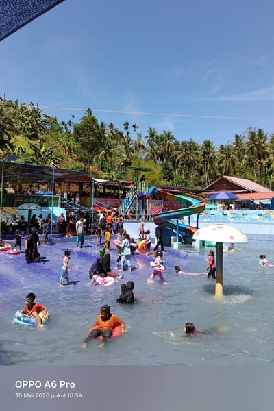 The image shows a swimming pool area in Palohseulimeng, Indonesia. A large group of people, mostly children, are swimming and playing in the water. Some are using inflatable rings. A colorful waterslide is located near the pool's edge. Some people are standing or sitting on the tiled deck surrounding the pool. There are buildings with roofs visible in the background, along with trees and vegetation. The sky is blue and clear.