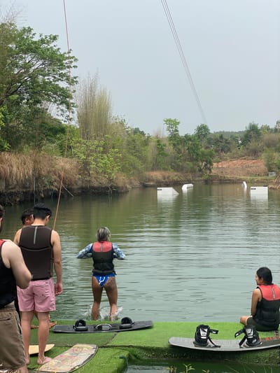 The image depicts a wakeboarding park located in Tambon Nam Phrae, Thailand. The scene is captured on a cloudy day. A body of water is visible, with wakeboarding obstacles placed within it. People are preparing to wakeboard on the water's edge. One person is already in the water, holding onto a wakeboard. On the side is artificial turf for people to stand on. Trees and other natural vegetation surround the water and the horizon shows a hilly terrain. There are cables for the wakeboarding equipment visible in the sky.