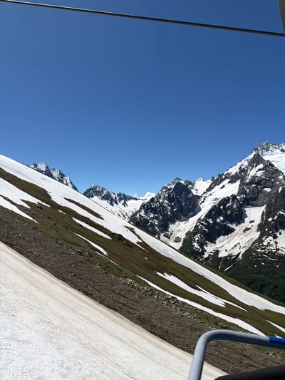 The image depicts a scenic mountain landscape under a clear blue sky. A thick steel cable stretches across the top of the frame, suggesting the presence of a cable car or ski lift. The foreground is dominated by a snow-covered slope, with patches of brown earth and green vegetation visible. This slope is part of a larger mountainside, with more snow-capped peaks in the distance. The mountains are rugged and rocky, with stark contrasts between the white snow and dark rock. A dense green forest is visible in the valley between the mountains. The scene appears to be from a high vantage point, possibly from the cable car itself, looking out over the vast mountainous terrain. The overall impression is one of natural beauty and grandeur, typical of a high-altitude alpine environment. The location is specified as Verkhne-Teberdinskoye Rural Settlement, Russia, suggesting a mountainous region within that administrative area.
