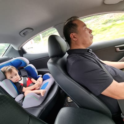 A man is driving a car, and a baby is in a car seat in the back. The baby is looking at the camera. The man is looking out the window. The car is on a road, and there are trees and grass outside. The image was taken in Gangnim-myeon, South Korea.