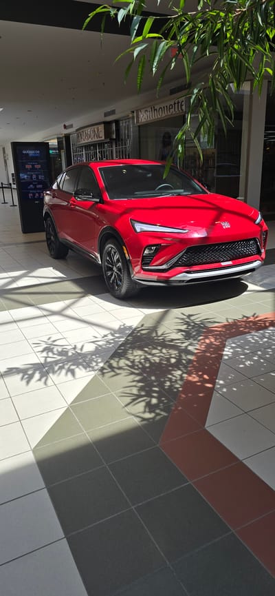 In Victoriaville, Canada, a vibrant red 2024 Buick Envista is displayed prominently in the interior of the Laurier Québec shopping mall. The vehicle is positioned on a checkered floor with geometric patterns of white, gray, and reddish-brown tiles. Sunlight streams in from above, casting dappled shadows of tree leaves across the car and the floor, suggesting an outdoor element is visible through the mall's architecture.

The car, a compact SUV, is the central subject, viewed from a slightly elevated, three-quarter front angle. Its sleek design, with sharp lines and a distinctive grille, is highlighted by the bright lighting. The vehicle's black wheels and tinted windows add a touch of modern contrast to its red exterior.

In the background, storefronts are visible, including "PENTAGONE" and "SB Silhouette," indicating retail establishments within the mall. A digital display board for "QUÉBECOR AFFICHAGE" is positioned to the left of the car, providing information on advertising services. The overall environment is that of a well-lit, modern commercial space, with architectural elements like ceiling structures and lighting fixtures suggesting an indoor setting designed to mimic natural light and bring in natural elements like the overhead foliage. No people are clearly depicted in the scene.