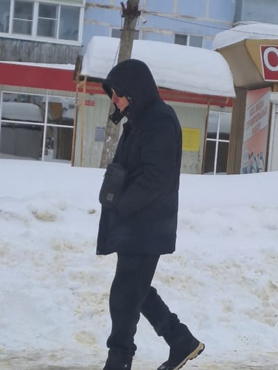 An individual is depicted walking from right to left on a snow-covered street in Sasovo, Russia. The person wears a black hooded puffer jacket with the hood up, dark sunglasses, black gloves, black pants, and black winter boots with a tan sole. A black shoulder bag is visible on their right hip. The ground is covered in white and discolored snow, indicating a winter environment. In the background, multiple buildings are visible, including one with a light blue upper facade, a wooden lower section, and windows, along with a red awning piled with snow. Adjacent to this, another commercial building or shop entrance features a yellow awning also covered in snow, and a red sign with a white circular logo (possibly an 'O'). A weathered utility pole stands between the individual and the background structures.
