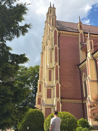 In the city of Гервяцкі сельскі Савет, Belarus, a person stands in front of a large, red-brick church with yellow architectural accents and ornate spires. The church's facade is detailed with arched windows and decorative banding. The person, wearing a light-colored t-shirt, is facing away from the camera and holding a mobile phone, suggesting they might be taking a picture or looking at something on the device. Lush green trees and manicured bushes surround the church, creating a natural, verdant setting. The sky above is partly cloudy, with patches of blue visible.