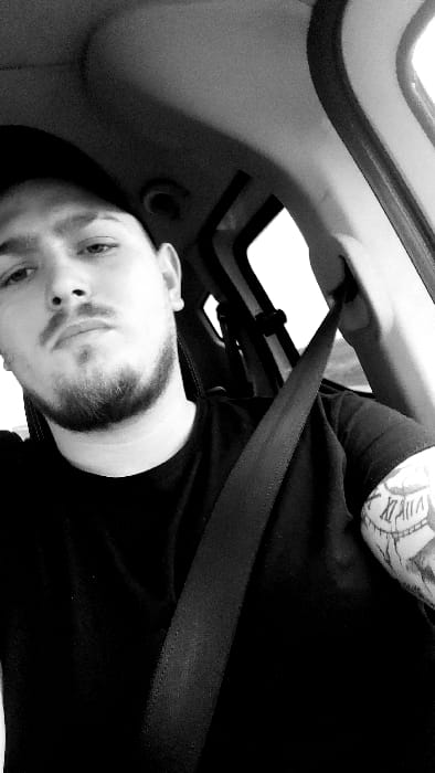 The image is a monochrome, vertically oriented selfie taken inside a motor vehicle, depicting a young adult male. He is seated, wearing a dark-colored t-shirt and a dark baseball cap, with a seatbelt diagonally crossing his chest from his right shoulder to his left hip. The individual has a short beard, mustache, and dark eyebrows, and is looking directly towards the camera with a neutral expression. A tattoo featuring Roman numerals (e.g., "XI II XII") and other patterns is visible on his left bicep/shoulder area. The background shows the interior of a car, including the headliner, parts of the vehicle's frame (A-pillar), and bright, overexposed windows suggesting daylight outside. The image is stated to be located in Senec, Slovakia.