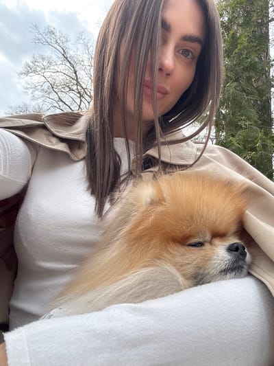 The photograph shows a person holding a Pomeranian dog. The person has long, dark hair, fair skin, and green eyes. They are wearing a white top and a beige jacket. The dog is mostly orange and is nestled in the person's arms, appearing to sleep.
Based on the surrounding environment, which includes trees and greenery, the location appears to be outdoors. The reference to Konstancin-Jeziorna, Poland, is not visually substantiated in this image. Without further information, pinpointing the specific location remains uncertain.