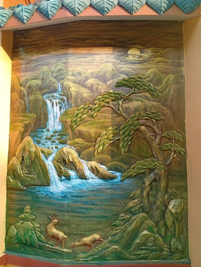 This is a vibrant, multi-layered relief sculpture mural, likely situated on an exterior wall, depicting an idyllic natural landscape. The scene features lush green mountains and cliffs, from which a series of waterfalls cascade into a winding river or stream, rendered in bright blues and whites, with visible rocks in its bed.
In the foreground, two golden-brown deer are present: one standing proudly with visible antlers, looking left, and another, smaller one, bent down as if grazing or drinking by the water's edge. A prominent, large, gnarled tree with stylized green and gold foliage dominates the right side of the mural, its branches extending over the water.
The background showcases distant, rolling mountains under a sky filled with artistic clouds, where a distinct golden moon is visible in the upper right. The color palette is rich, with varying shades of green, earthy browns, and deep blues, enhanced by golden highlights on the raised elements like the deer, tree leaves, and some rock faces, suggesting either dawn/dusk or a stylized mystical light.
Above the mural, a decorative roofline is visible, adorned with light blue-green, leaf-shaped or scale-like tiles, hinting at traditional East Asian or Vietnamese architecture, consistent with the Phú An, Vietnam location context. The overall impression is one of tranquility and natural beauty, rendered with striking three-dimensional texture.