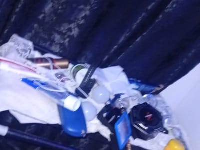 The image depicts a cluttered surface with various objects, including what appears to be a blue container, white cloths, a black electronic device resembling a speaker, and other unidentifiable items. The background is a dark, textured fabric. The scene appears to be indoors. No specific location within Pantinapu, Nigeria is discernible from the image.