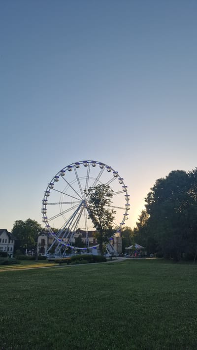 This image shows a large Ferris wheel in a park in Keszthely, Hungary, likely during the late afternoon or early evening. The sky is clear and a pale blue. The Ferris wheel is the dominant subject, its white structure and numerous gondolas illuminated with blue lights. Behind the Ferris wheel, the setting sun casts a warm glow, visible through the trees. The park itself features a well-maintained grassy lawn in the foreground, with a path and some benches. In the background, several buildings with traditional European architecture can be seen, as well as lush green trees. A small carousel can also be spotted to the right of the Ferris wheel. There are no people visible in the image.
