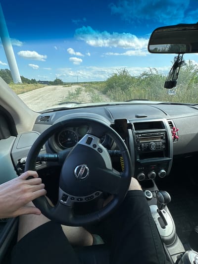 This media file shows the interior of a car, likely a Nissan, with a person's hands on the steering wheel. The scene is set in a car with a dashboard featuring a radio, air vents, and gear shift. Outside the car, a bright blue sky with scattered white clouds is visible through the windshield and side window, suggesting a clear, sunny day. The landscape outside appears rural, with dry grass and fields. The time of day is likely midday due to the brightness of the sun. A hand is visible holding the steering wheel, indicating the car is being driven or is about to be driven. There are no strong emotional cues visible, but the scene suggests a casual, everyday activity. The image is oriented vertically, with the top of the car interior appearing on the left side of the frame. Text on the steering wheel clearly reads "NISSAN". Other visible text includes radio display information and what appears to be a "NO" or "OFF" label on a control.