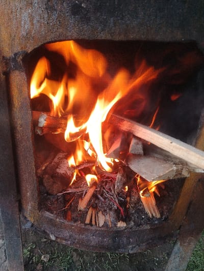 A close-up photograph taken in Nurli zamin, Uzbekistan, depicts an active fire contained within a weathered, dark metallic combustion chamber or stove. The chamber, likely cylindrical or barrel-shaped, features an irregular, roughly arched opening at the front, through which bright orange and yellow flames are actively burning. Inside, various pieces of wood, including logs, kindling, and splinters, are visible as fuel. One light-colored wooden plank extends from the upper right, diagonally into the flames. Below the burning wood, a bed of ash, charred material, and glowing red embers is present. The internal surfaces of the metal structure are heavily sooted and show signs of corrosion. The base of the metallic chamber rests directly on an outdoor ground surface consisting of green grass and patches of dry soil. No individuals, animals, or additional significant objects are depicted.