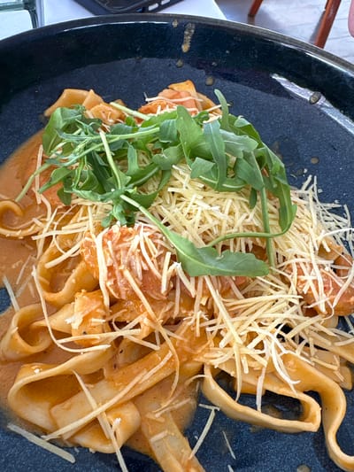 A close-up, overhead shot shows a dark, textured plate filled with fettuccine pasta coated in a creamy, orange sauce. The pasta is topped with a generous portion of shredded cheese and a small pile of fresh, green arugula leaves. The dish appears to be served in a restaurant setting, with hints of a white tablecloth and wooden furniture visible in the blurred background. The lighting suggests an indoor environment, possibly during the day.
