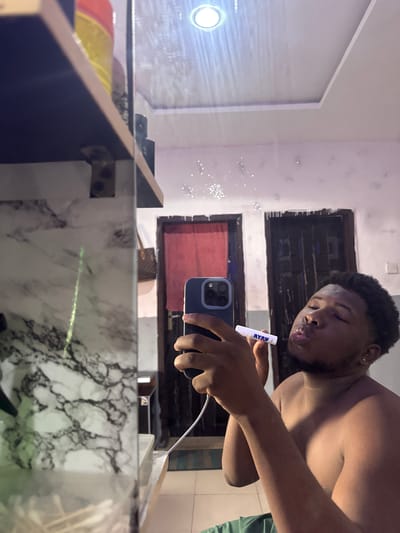 A shirtless Black male in Gbongan, Nigeria holds a blue mobile phone in his left hand while blowing bubbles with a white bubble wand held in his right. He is likely taking a selfie in front of a bathroom mirror. The bathroom has dark wooden shelves and a marble countertop. A small white light is fixed above the mirror. Two dark wooden windows with visible shutters sit behind the man's reflection in the mirror.