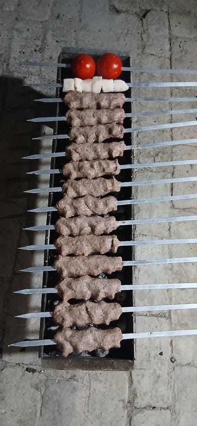 In the city of Andijani, Uzbekistan, a barbecue grill is laden with numerous skewers of food. The grill, made of dark metal, rests on a surface that appears to be made of rough, light-colored bricks. The background consists of a brick wall.
At the top of the grill, two ripe, red tomatoes are skewered. Below them are several pieces of white, cubed food, possibly chicken or fish. The majority of the skewers are filled with what appears to be ground meat, shaped into long, cylindrical kofta or kebabs. These meat skewers are arranged in rows, with the tips of the metal skewers extending outward. The meat is a reddish-brown color and has a textured surface. The scene is illuminated by a light source that casts subtle shadows. The overall impression is one of food preparation for a meal, likely outdoors given the presence of the grill and brickwork.
