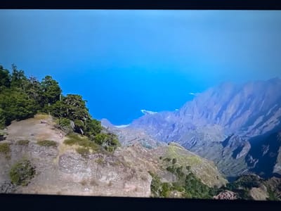 The image shows a panoramic view of a rugged, mountainous coastline under a clear blue sky. In the foreground, a steep, rocky slope is covered with sparse vegetation and a few trees. A small figure, possibly a hiker, can be seen on a dirt path traversing the slope. Further in the distance, the terrain descends into deep valleys and ridges, revealing layers of rock formations that lead to the sea. The ocean is a vibrant blue, meeting the land at a hazy horizon. There is no indication of the city of Ləcət, Azerbaijan, in this image.