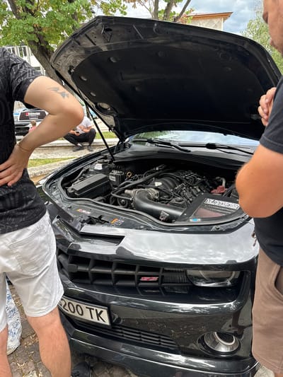 In Hadzhidimovo, Bulgaria, a black Chevrolet Camaro with its hood open is parked outdoors. The engine is visible, and modifications such as an aftermarket air intake are present. Two individuals are standing near the car; one is wearing a black t-shirt and white shorts, and the other is wearing a black t-shirt and brown shorts. A third person is partially visible in the background, sitting on steps. The environment appears to be a residential or public area with trees and buildings visible. A license plate with Cyrillic characters and the designation "200 TK" is affixed to the front of the car.