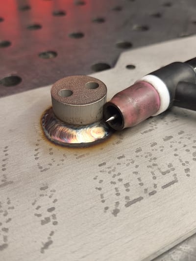 The image shows a close-up view of a metal surface with a TIG welding torch positioned next to a small, cylindrical metal object that has two holes in it. The object is welded to the surface, and the weld shows iridescent heat discoloration, indicating it is a fresh weld. The metal surface itself appears to be a worktable or a piece of equipment with a pattern of small, dark spots and a metallic sheen. In the background, a red surface with circular indentations is visible, possibly another part of the work area or equipment. There are no people visible in the image. The scene is likely from a workshop or manufacturing environment where metal fabrication is taking place. The specific location in Bad Honnef, Germany, is not discernible from the image itself, as there are no identifying landmarks or contextual clues.