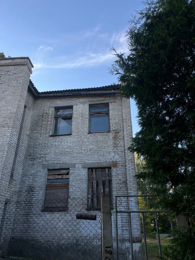 The image depicts the exterior of an old, light-colored brick building under a clear blue sky with wispy clouds. A large, dense green tree with fine needles partially obscures the upper portion of the building and the sky. The building appears weathered, with visible mortar lines and some staining on the brickwork.

There are several windows visible. Two rectangular windows are intact with dark frames and reflect the sky. Another opening has wooden planks haphazardly nailed across it, suggesting disrepair. A smaller square opening is also covered with wooden planks. Above these, a larger opening is boarded up with rough wood. A metal pipe protrudes from the brick wall near one of the boarded windows.

A chain-link fence runs along the front of the building, with some overgrowth visible beyond it. A metal railing system is also present near the fence. On a horizontal concrete beam or structure in front of the fence, the number "182" is written in black.

The scene suggests an outdoor setting, likely in a rural or semi-abandoned area. The time of day appears to be daytime, given the clear sky and lighting. There are no people visible in the image. The overall mood conveyed is one of neglect and age. The location context provided is Астроміцкі сельскі Савет, Belarus.