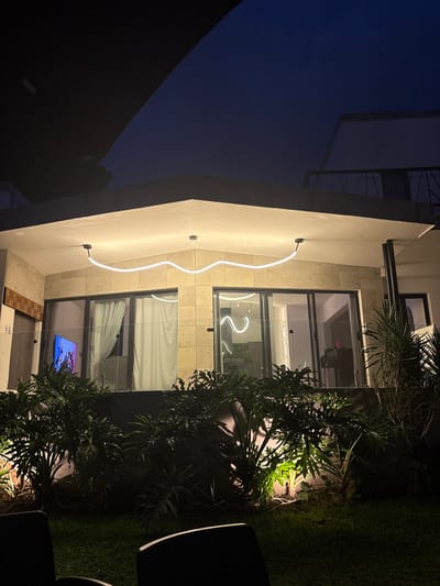 The image shows an exterior shot of a modern building in Tres de Mayo, Mexico, taken at dusk or night. The building has a white facade with stone accents. A decorative neon light fixture, shaped like a wavy line, is mounted vertically on the facade. Large windows with dark frames reflect interior lighting. Behind the windows, the faint outline of furniture can be seen, as well as a TV screen. In the foreground, dense foliage and landscaping are illuminated by ground lights. Part of what appears to be outdoor furniture can be seen. The sky above is a deep blue, suggesting the photo was taken shortly after sunset. The image is rotated and appears to be taken from a low angle.