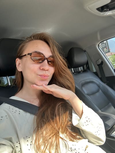 A woman is seated in the driver's seat of a car, wearing a seatbelt, a white shirt with a patterned cut-out design, and brown-tinted glasses. Her right hand is near her face. The interior of the car is visible, including the grey seats and car fixtures. Through the car window, part of a building with text written in Russian is visible. Based on the text, the location is inferred to be in Uva, Russia.