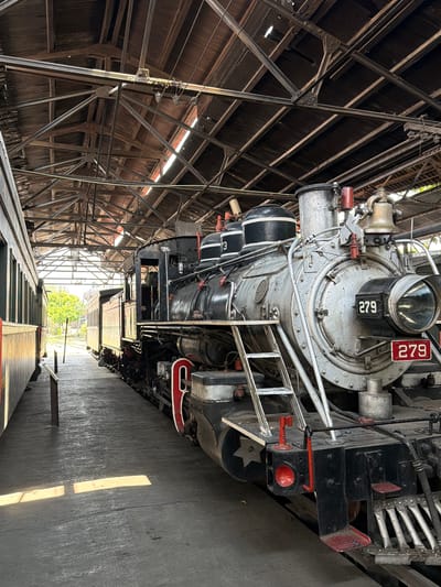 The image shows a vintage steam locomotive, number 279, likely inside a maintenance building in Cuautla, Mexico. The locomotive is silver and black with red accents. It has a large headlight and a number plate with "279" displayed. The building has a wooden and metal framework roof. The locomotive is parked on train tracks with other train cars. Sunlight is visible at the end of the maintenance building.