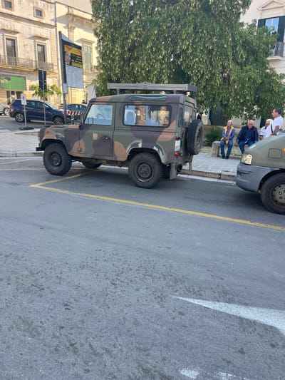 The image shows a military vehicle parked on a street in Galatina, Italy. The vehicle is camouflage-colored and has a spare tire mounted on the back. Several people are seated and standing nearby. The location is in the city center and is bordered by buildings with advertisements on them, and trees. There are also other cars parked along the street. The street surface is asphalt, marked with yellow and white lines for parking spaces and traffic direction.