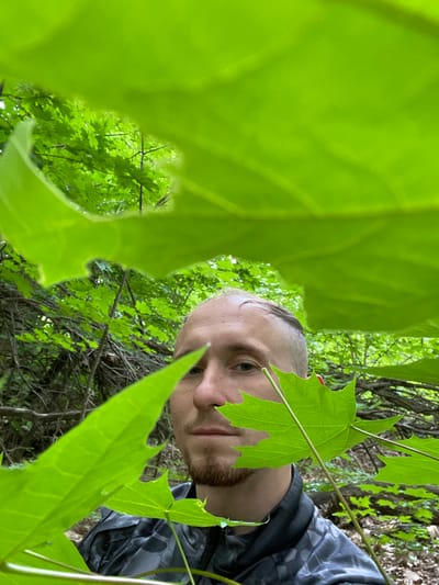 A person is in a forest environment. The person is partially obscured by large green leaves in the foreground, with only their face, neck, and upper torso visible. They have a light complexion, short-cropped hair, and a short beard. They are wearing a dark, patterned jacket. The background consists of dense green foliage, suggesting a woodland or park area. There is no information available that places this image in the city of Тияпино, Russia.