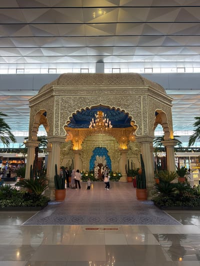 This media file captures a grand indoor space, likely an airport terminal or large commercial complex in Pajang, Indonesia, during daytime. The central subject is a large, ornate golden pavilion or archway, richly decorated with intricate carvings, arches, and a domed ceiling. Inside the structure, a blue patterned backdrop and a sparkling chandelier are prominently displayed. The pavilion is flanked by decorative columns and an array of potted plants, including tall cacti and leafy green foliage.
Several people are visible in the scene: a small group stands near the ornate structure, observing it, while another person walks away on the right. The floor leading up to the pavilion features a patterned rug, transitioning to a highly reflective, light-colored tiled floor on the right, which shows clear reflections of the architecture and surrounding plants.
The background showcases a modern building interior with a high, geometrically patterned ceiling and large windows that allow ample natural light. In the distance, various shops are discernible, with visible text including "OLDTOWN" and "MARRIOTT" on their signs. A red sign on the floor, partially visible on the right, reads "NO CART ALLOWED," indicating specific regulations for the area. The scene blends modern architectural elements with opulent, traditional-inspired design, creating a luxurious and spacious environment.