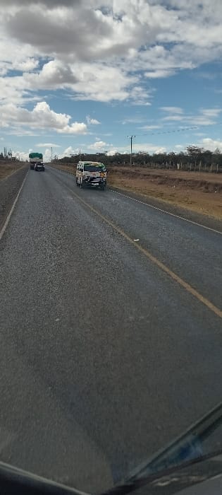 A road stretches into the distance under a cloudy sky, with a vehicle ahead of the camera. A colorful bus rides on the left side. The road is paved, with yellow dividing lines and a grassy shoulder on the right. Utility poles line the horizon on the left. The scene is taken from inside a vehicle, with a portion of the car's interior visible on the right side.