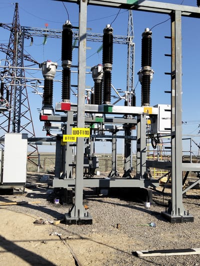 The image shows an outdoor electrical substation in Батыр ауылдық округі, Kazakhstan. The main focus is a metal structure supporting several large, black, ribbed insulators, each topped with a metal cap and connected by wires. These insulators are arranged in a row. To the left, a white rectangular electrical enclosure is visible. Yellow signs with black Cyrillic text are affixed to the metal structure, with one sign clearly reading "ШО-Т-1-110" and another below it reading "В-Т-1-110". In the background, a large metal tower with a lattice structure supporting overhead power lines stands against a clear blue sky. The ground around the substation is covered with gravel and some dirt, with a few scattered construction materials like wooden planks and a white paint can. The overall scene suggests an active or recently constructed part of an electrical power transmission or distribution system. There are no people visible in the image.