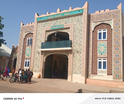The image captures the Emir's Palace in Wase, Nigeria, under a bright sky. The palace entrance is decorated with intricate geometric patterns in shades of beige, brown, blue, green, and yellow. A turquoise sign reads "EMIR'S PALACE WASE" and "SARKIN DUTSE" above the arched entrance. The building facade has two rectangular windows with blue bars and white frames on the right side. The windows on the left side of the building vary in size and shape. People are gathered near the palace entrance, with some sitting inside the archway and others standing outside. A man is on a motorcycle on the street in front of the palace, with people near it. The environment is a paved street, and a low, white-roofed building is visible to the left.