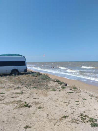 A white bus is parked on a sandy beach in Dolzhanskaya, Russia, with sparse vegetation. The sea is in the background, with waves and a clear blue sky. There are a few people in the water and a red kite in the sky.