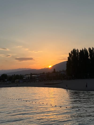This is a vertical, eye-level, medium shot of a sunset over a body of water, likely a lake, with a beach and some trees in the background. The sun is low on the horizon, casting a warm orange and yellow glow across the sky and reflecting on the water. The water's surface is rippled, creating a textured pattern of light and shadow. A line of buoys stretches across the water. On the beach, a few people are visible, some walking and one standing near the water's edge. The beach appears to be lined with trees and some structures, possibly part of a resort or village. The overall mood is peaceful and serene, suggesting a calm evening at a lakeside location. The time of day is clearly sunset, and the weather appears to be clear. The location cues of trees and mountains in the distance, along with the presence of a beach and body of water, are consistent with a lakeside setting. No visible text is present in the image.
