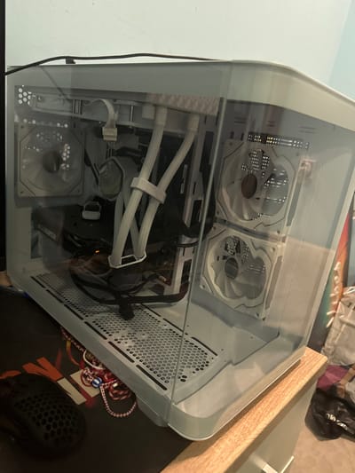 The image depicts an indoor scene featuring a white personal computer (PC) case with a transparent side panel, positioned on a light-colored wooden surface. The PC case contains various internal components. Visible internal elements include a white All-In-One (AIO) liquid cooler with a pump block and two white hoses, which are secured by a white plastic cable management clip. Three white cooling fans are present: one at the rear of the case and two mounted vertically on the right side. A dark-colored graphics card is installed horizontally near the bottom-center, exhibiting a small orange light or reflection. Several black cables are routed throughout the interior, with some resting loosely on the perforated bottom panel of the case. A white cable bundle is connected at the top-left interior.
To the immediate left of the PC case, a black gaming mouse with a honeycomb shell design is visible, resting on a black mousepad featuring partially visible red and orange graphics. The background consists of a light blue wall. On the far right, blurred items, possibly clothing or textiles, are partially discernible.
The image provides no visual information to confirm the specified location in the city of Topolchane, Bulgaria.