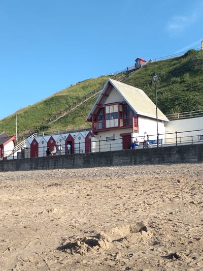 The image shows a view of the beach and cliffside in Saltburn-by-the-Sea, United Kingdom. In the foreground, there is a sandy beach with tire tracks visible. A low stone wall separates the beach from a promenade area. Behind the wall are a row of red and white beach huts. Further back, a white building with a red-trimmed roof and windows sits on the lower part of a grassy, green hillside. A set of stairs with lights runs up the hillside, and a small red building with a pointed roof is visible near the top of the hill. The sky is clear and blue. Several people are visible on the promenade, some standing near the beach huts and others on benches. One person is sitting on a bench and another is standing near them. Another person is walking away from the camera on the promenade.