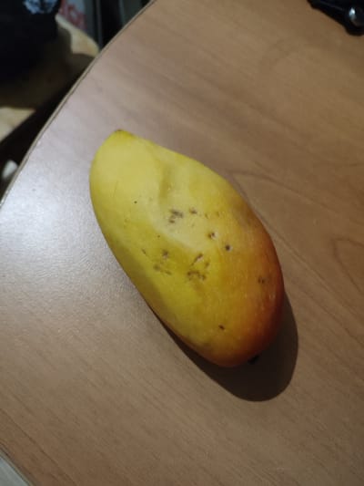 A single, yellow-orange mango rests on a light brown wooden surface. The mango is slightly oblong with some dark spots visible on its skin. The lighting suggests an indoor environment, possibly a table. The image does not contain any elements that would indicate a specific location in Lisakovsk, Kazakhstan.