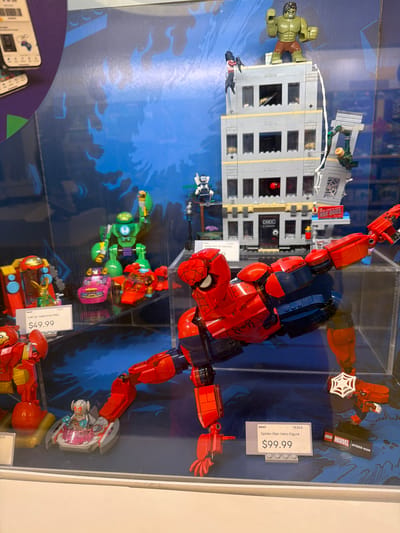 The image displays a retail store window in Mission Viejo, United States of America, showcasing various LEGO Marvel superhero merchandise. The primary focus is a large LEGO Spider-Man figure in a dynamic pose, positioned prominently in the foreground. Behind Spider-Man, a LEGO building is depicted with several figures including the Hulk, Spider-Man, and other characters engaged in what appears to be a battle scene. To the left, smaller LEGO sets are visible, including a green mech suit, a pink vehicle, and a red vehicle with a Loki figure. A price tag for "Loki vs. Team Iron Man" is visible, indicating a price of $49.99. Further to the right, a price tag for "Spider-Man Hero Figure" is displayed with a price of $99.99, alongside a LEGO Marvel Spider-Man display. The background features a blue, comic-book style graphic with flame-like designs. The lighting suggests an indoor retail environment.