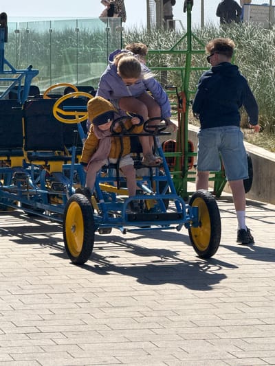 This image captures an outdoor scene in Middelkerke, Belgium, likely a recreational area or promenade. The primary subjects are several people and a large multi-person pedal kart.
In the foreground and mid-ground, a blue and yellow pedal kart is prominent. Seated on the kart are at least four individuals: an adult in a purple jacket, another adult (partially obscured), and two children. One child, wearing a yellow/brown coat and a striped shirt, is holding a yellow steering wheel, appearing engaged in steering. Another child is partially visible next to them.
Standing to the left of the kart is a young man with light brown hair, wearing a dark jacket, light blue denim shorts, white socks, and dark shoes. He is also wearing sunglasses and is looking down, possibly at the people on the kart or the ground.
The activity appears to be recreational, with the group either preparing for a ride on the pedal kart or having just completed one.
Notable details include the bright sunlight, which casts long, distinct shadows of the people and the kart onto the light grey paved ground, suggesting it is either late morning or early afternoon on a clear day. In the background, there is green vegetation resembling dune grass and a glass fence, consistent with a coastal or resort setting like Middelkerke. A dark bag or jacket hangs on a green metal fence section in the upper left background. No discernible text is visible in the image. The overall atmosphere appears casual and relaxed.