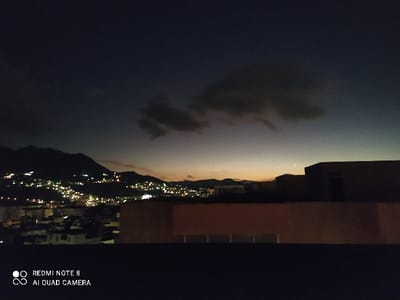 Night scene depicts a cityscape nestled against mountains, illuminated by scattered lights resembling stars. Above, a dark sky is partially obscured by clouds with a subtle gradient of sunset glow on the horizon. The foreground features a rooftop with structures, silhouetted against the dusk. The camera, identified as Redmi Note 8 AI Quad Camera, captures a low-light perspective.