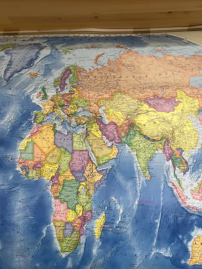 A physical and political world map, predominantly in Cyrillic script, is displayed in an indoor setting, likely affixed to a wall or large surface, with a wooden border visible on the left side. The image is rotated 90 degrees clockwise, showing the map with Europe and Africa positioned on the left, Asia on the right, and the Arctic region at the top.

The map illustrates geographical features and political boundaries using varied colors. Landmasses are depicted in different hues to distinguish countries, such as the large orange expanse of Russia ("РОССИЙСКАЯ"), the yellow regions of China ("КИТАЙ") and Mongolia ("МОНГОЛИЯ"), and the green of India ("ИНДИЯ"). The continent of Africa is prominently featured in diverse colors, showing countries like Algeria ("АЛЖИР"), Libya ("ЛИВИЯ"), Egypt ("ЕГИПЕТ"), Sudan ("СУДАН"), South Sudan ("ЮЖНЫЙ СУДАН"), Angola ("АНГОЛА"), Namibia ("НАМИБИЯ"), Botswana ("БОТСВАНА"), and the South African Republic ("ЮЖНО-АФРИКАНСКАЯ РЕСПУБЛИКА").

Oceans are rendered in shades of blue, with bathymetric details indicating underwater topography. Visible ocean labels include "ИНДИЙСКИЙ ОКЕАН" (Indian Ocean), "БАРЕНЦЕВО МОРЕ" (Barents Sea), "ВОСТОЧНО-СИБИРСКОЕ МОРЕ" (East Siberian Sea), and parts of the "АТЛАНТИЧЕСКИЙ ОКЕАН" (Atlantic Ocean). Greenland ("ГРЕНЛАНДИЯ") is visible in the top left, and Madagascar ("МАДАГАСКАР") is off the coast of southeastern Africa. The map provides a detailed overview of the Eastern Hemisphere, with clear labeling of countries, major cities, and bodies of water in Russian. No activity is taking place; it's a static image of the map.