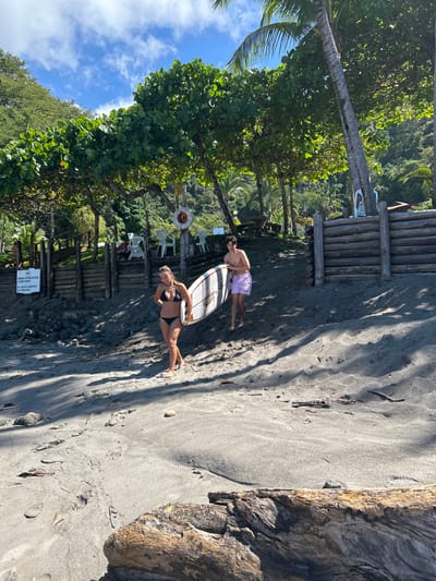 The image captures a sunny daytime scene on the dark sand beach of Playa Herradura, Costa Rica. Two individuals are featured, standing near the treeline. In the foreground, a female in a black bikini stands with a white surfboard tucked under her left arm, looking towards the right. Behind her and slightly to the left, a male wearing light-colored swim shorts stands with his hands on his hips.
The beach features dark, possibly volcanic sand, with visible footprints and scattered small rocks. To the right, a large, weathered piece of driftwood or a log lies partially buried in the sand. Behind the people, a rustic wooden platform or walkway is visible, furnished with several white plastic chairs. A red and white life preserver ring hangs from a post nearby. Lush, vibrant green tropical vegetation, including palm trees, borders the beach, creating a shaded area and extending into heavily forested hills in the background. The upper left corner shows a bright blue sky with sparse white clouds.
Long, distinct shadows cast by the trees and individuals indicate bright sunshine, suggesting it is likely late morning or early afternoon. A white sign with black text is partially visible near the wooden structure at the bottom left, displaying text including "PROHIBIDO EL PASO" (passage prohibited) and other unreadable words, possibly relating to rules like no littering. The overall atmosphere suggests a relaxed, tropical beach environment conducive to surfing.