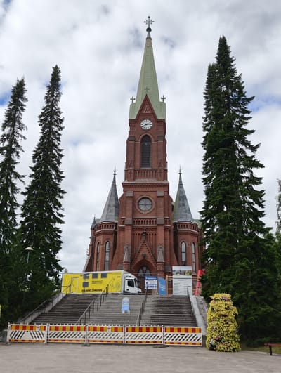 The image shows a red brick church with a tall green spire and a clock on its tower, located in Mikkeli, Finland. Tall evergreen trees flank the church. In front of the church are a set of stairs leading up to the entrance. A yellow construction trailer with the name "HAMMELMANN" is parked on the stairs. Safety barriers are in place at the bottom of the stairs. There are informational posters and signs around the construction area. A decorated bush with yellow flowers is situated on the right side of the image. The sky is partly cloudy.