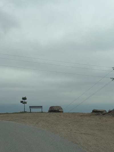 A landscape featuring an overcast sky, power lines, a rocky hillside, and a road. A bench and a small tree stand at the edge of the hill, near the road. The image might be taken in a rural or semi-rural area, potentially near Leshten, Bulgaria.