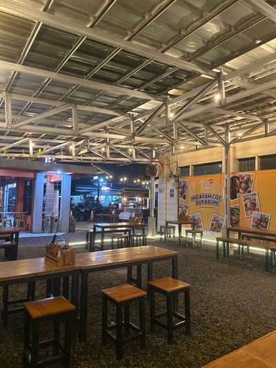 The image displays an illuminated outdoor or semi-outdoor dining area at night, located in Kecamatan Cisaat, Indonesia. The ground is covered with light-colored gravel, and numerous dining sets consisting of tall, narrow wooden tables with black metal frames and matching backless stools are arranged throughout the space.
On the left, a modern building with a facade of reflective panels is partially visible, overlaid by a prominent metal truss structure that supports the overhead space. This structure is adorned with multiple string lights, providing warm ambient illumination. A small plant with vines can be seen climbing a support column.
On the right side of the scene, a food stall or concession stand is marked by bright yellow banners. One banner clearly features the text "MIE AYAM COE SUKABUMI" along with appetizing images of various noodle dishes, indicating the specialty offered. Another yellow banner also displays food imagery and includes a phone number: "0896 7744 3129".
In the background, beyond the immediate dining area, other brightly lit establishments or parts of a street are visible, along with a few indistinct figures of people and parked motorbikes, suggesting a busy environment. A spherical convex mirror hangs from the ceiling structure. The time of day is clearly night, emphasized by the reliance on artificial lighting to brighten the setting. The overall atmosphere appears casual and inviting, typical of a lively outdoor eatery or food court.
