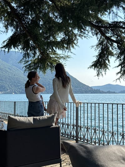 Two young women stand on a paved terrace overlooking a large body of water, likely Lake Como, given the mountainous backdrop characteristic of the region. The woman on the left is facing left and speaking, her hair pulled back in a ponytail, wearing a white tank top and denim shorts. The woman on the right faces away from the camera, gazing out at the water, her hair long and dark, wearing a cream-colored knitted sweater and a floral skirt. They are both leaning on a decorative black metal railing that separates the terrace from the water. In the foreground, dark grey outdoor seating furniture with large grey cushions is visible. The water is a vibrant blue, reflecting the clear sky and distant green, tree-covered mountains that frame the lake. Pine branches overhang the scene from above, casting dappled shadows. The overall ambiance is serene and picturesque.